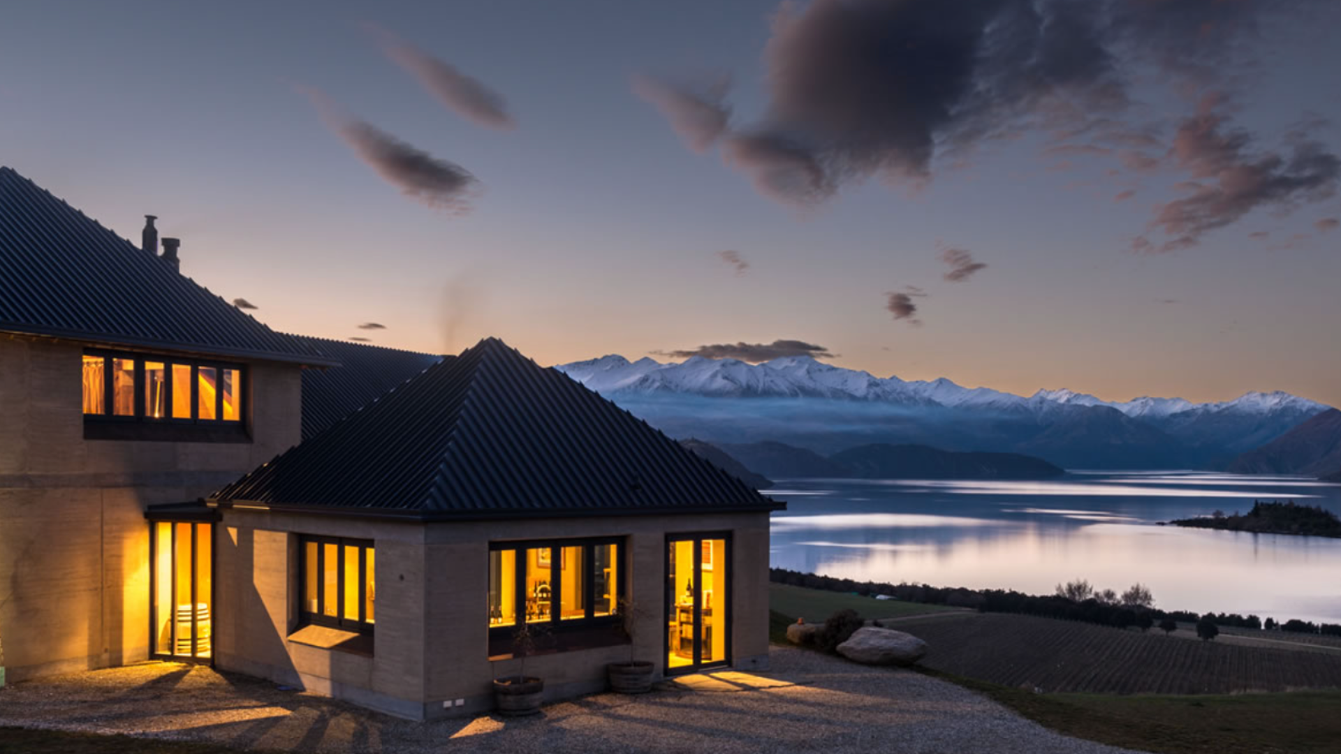 Rippon Vineyard building at dusk with warm lights, overlooking Lake Wanaka and the snow-capped Southern Alps